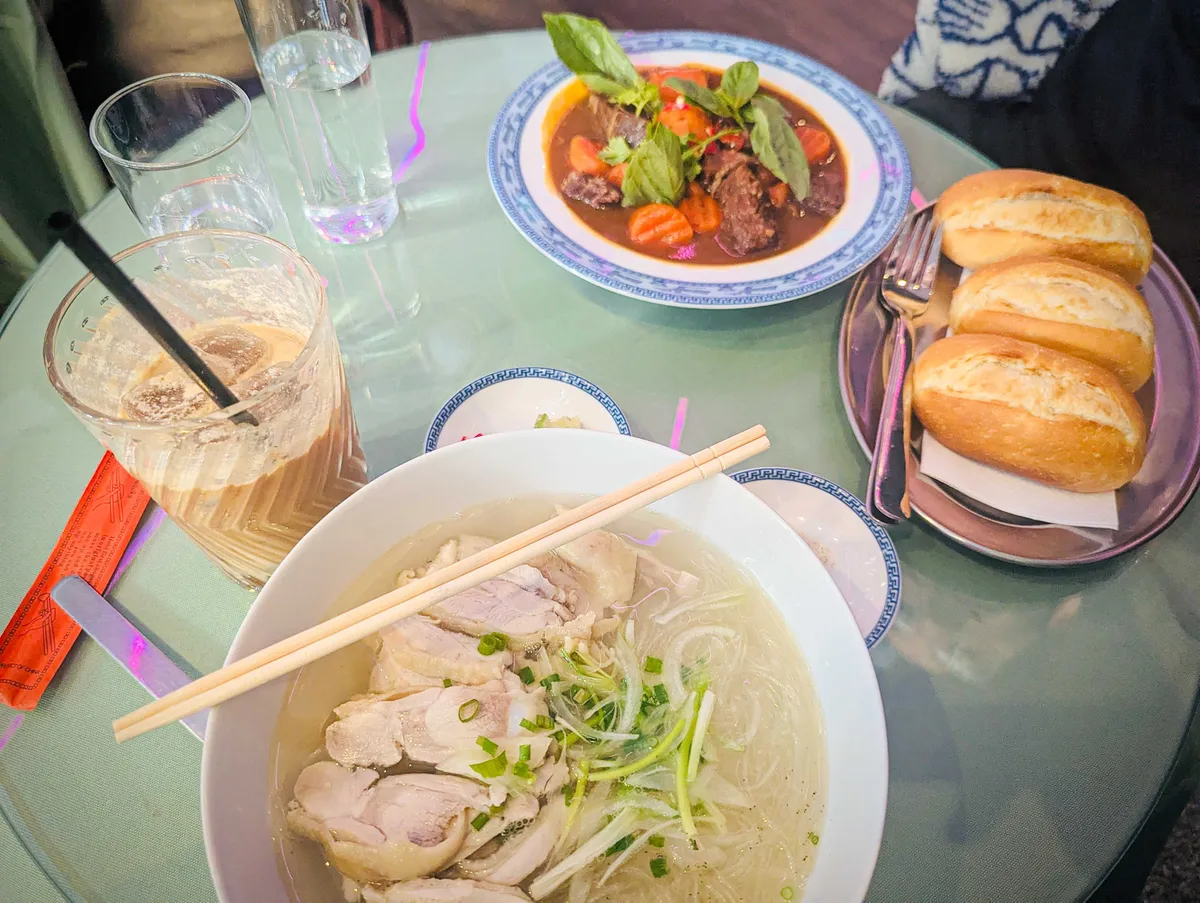 A bowl of chicken soup with glass noodles paired with a half-filled glass of an iced latte. Towards the back are 3 small baguette rolls and a plate of beef stew with carrots.