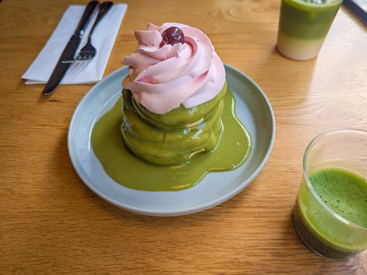 A small but tall stack of thick pancakes, covered in green sauce. A generous dollop of pink whipped cream is caked on top, with a cherry in the center.