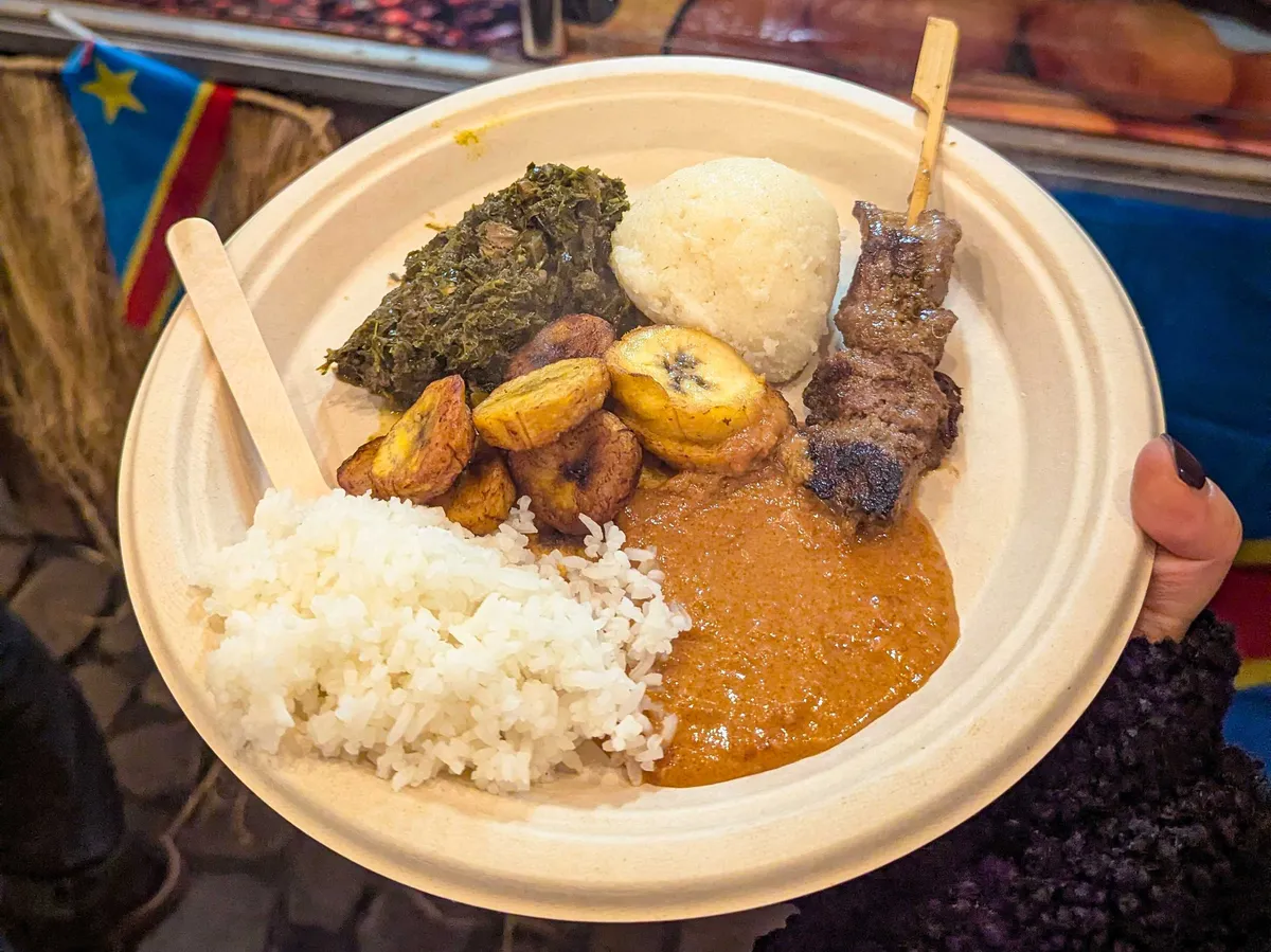 Peige holds a plate consisting of rice, fried bananas, cooked spinach, fufu, peanut sauce, and a meat skewer.