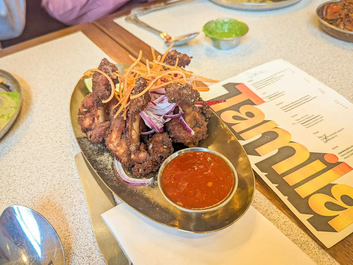 A stack of small chicken drumsticks on top of a gold plate that have been marinated and coated generously with black pepper. They've been topped with sliced carrots and red onions. There's a small dish of chili sauce on the side.