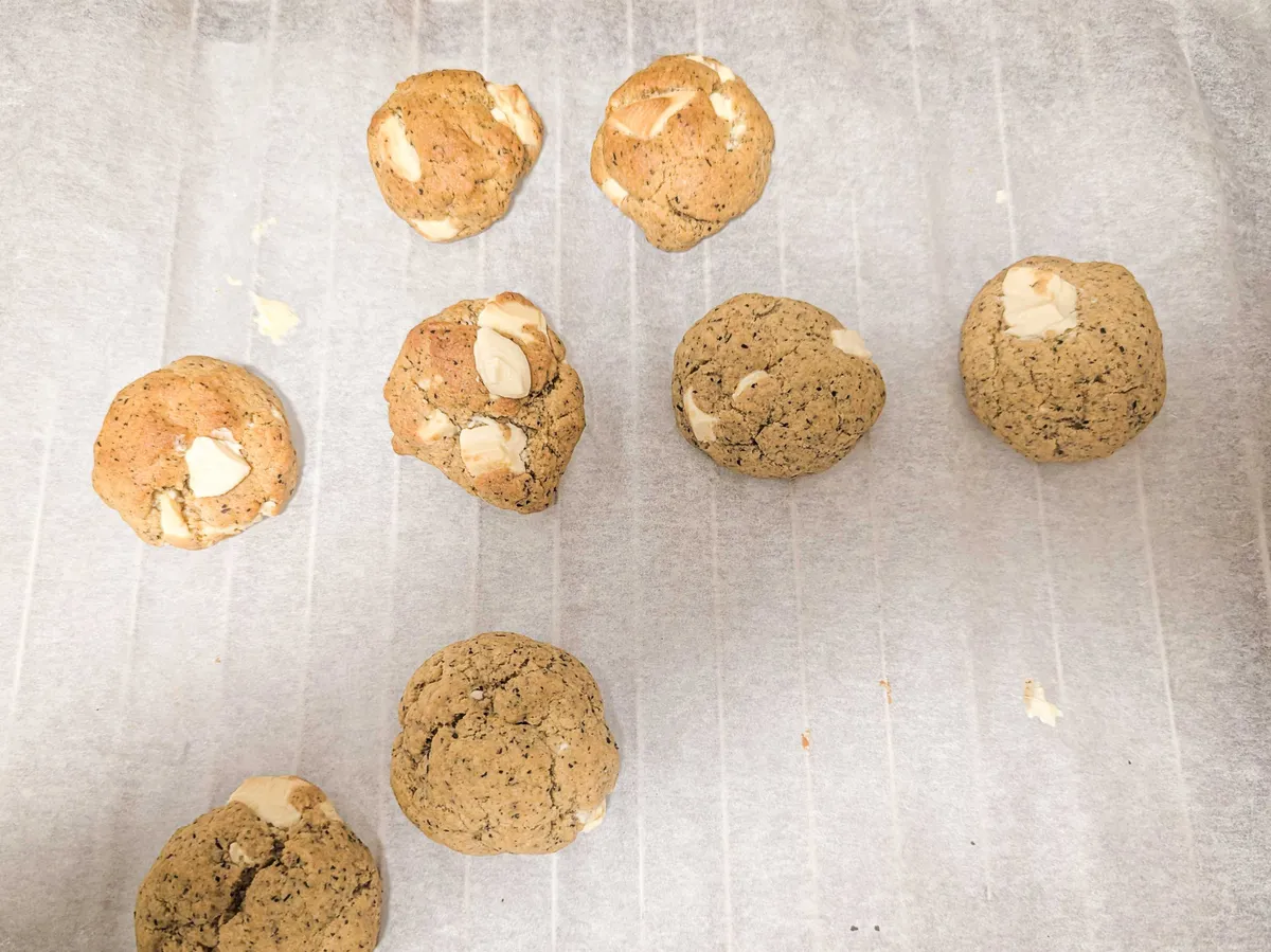8 cookies haphazardly displayed on a baking sheet. They are round and look quite firm, like scones. There are chunks of white chocolate bursting from some. All have a dotted pattern, like poppyseeds.