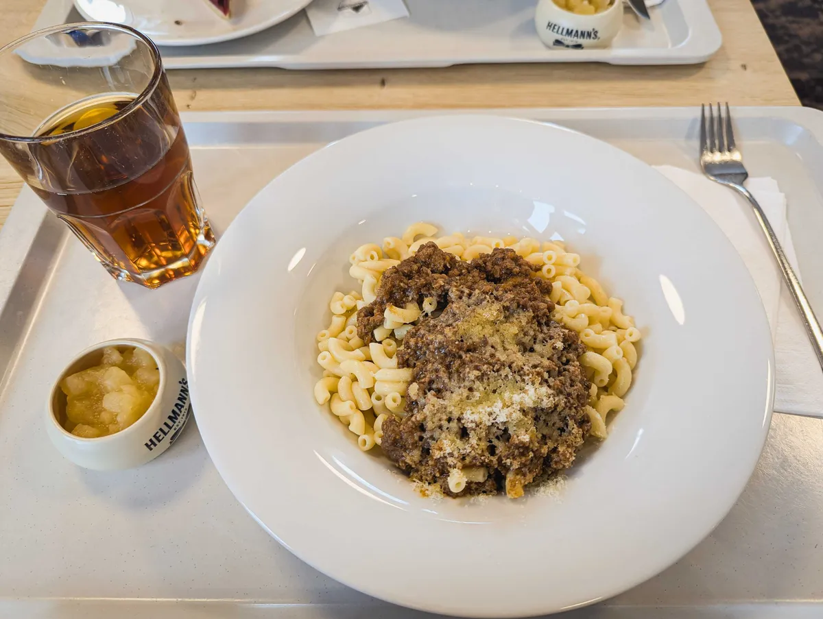 A dish of macaroni smothered in meat sauce with cheese sprinkled on top. A small dish of chunky apple sauce is on the side, and there's a glass of iced tea.