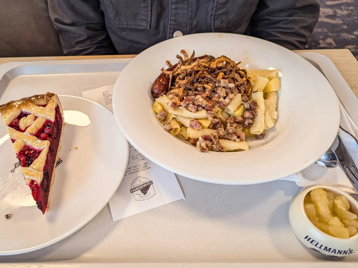 A dish of macaroni with a creamy bacon sauce topped with fried onions and one sausage. There's a small dish of apple sauce on the side, and a slice of berry pie.