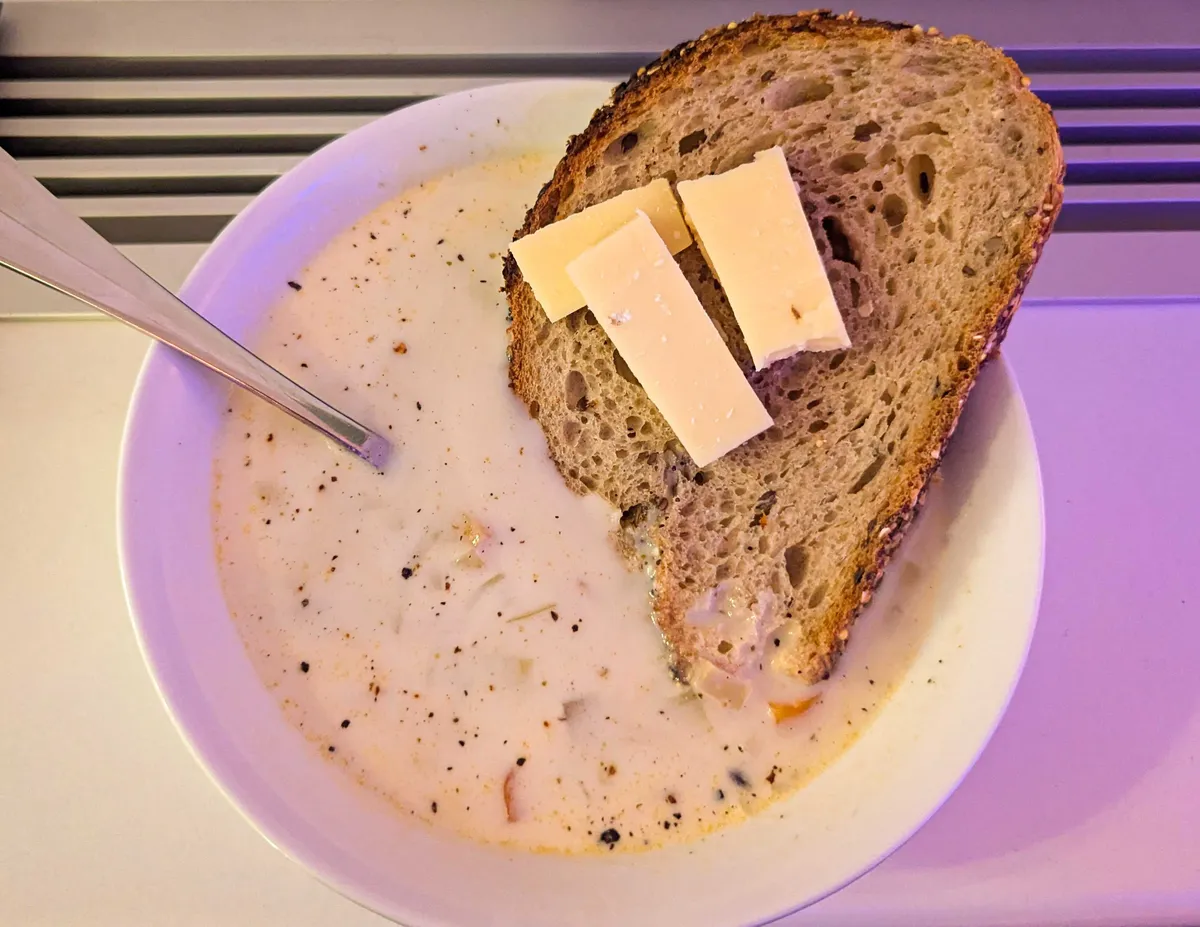 A bowl of light colored soup with bits of vegetables and black pepper. A slice of bread is dipped into the soup, with three small slices of cheese sitting on the bread.