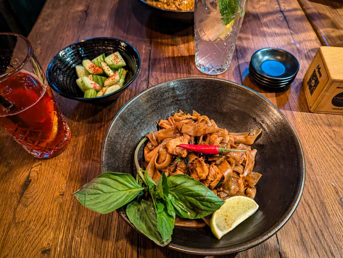 A plate of wide rice noodles, stir-fried with soy sauce, chicken, and chilli. There is a sprig of Thai basil and a wedge of lemon. To the left is a glass of hibiscus iced tea and a small bowl of cucumber salad dusted with chilli flakes.