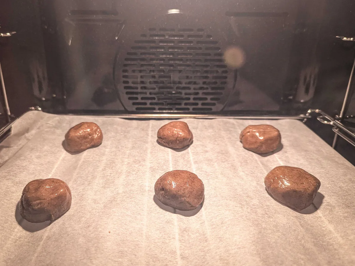 Two rows of 3 dark brown dough balls inside the oven on top of baking paper.