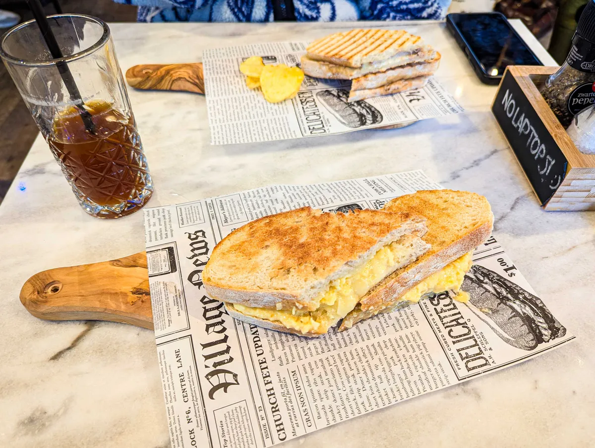 A sliced toastie on sourdough bread, served on top of a wooden cheeseboard lined with a fake newspaper. An iced Americano in a tall glass with a straw is to the side. Daniel's toastie and chips are in the background.