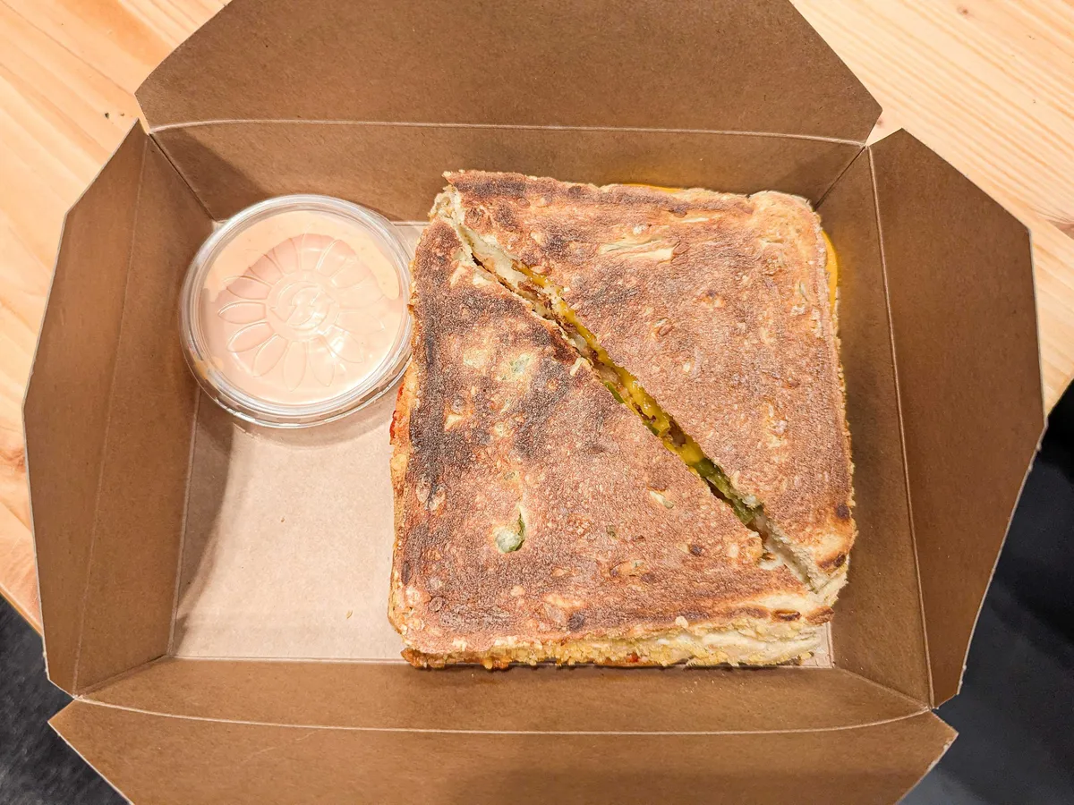 An open to-go box with a square toasted sandwich that's been cut diagonally in half, and a small plastic tub of light orange sauce.