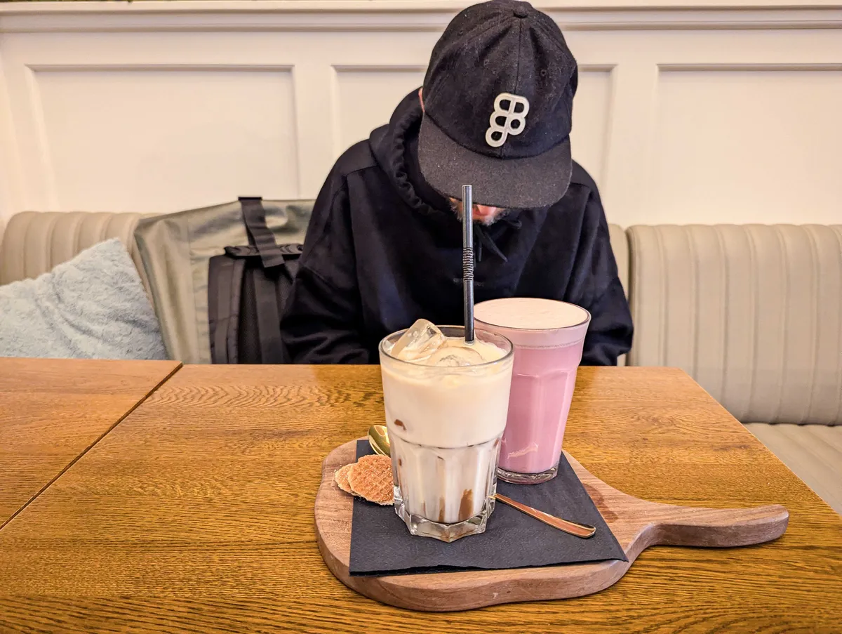 A cream colored drink and a pink colored drink served together on a wooden cutting board, with mini stroopwafels on the side.