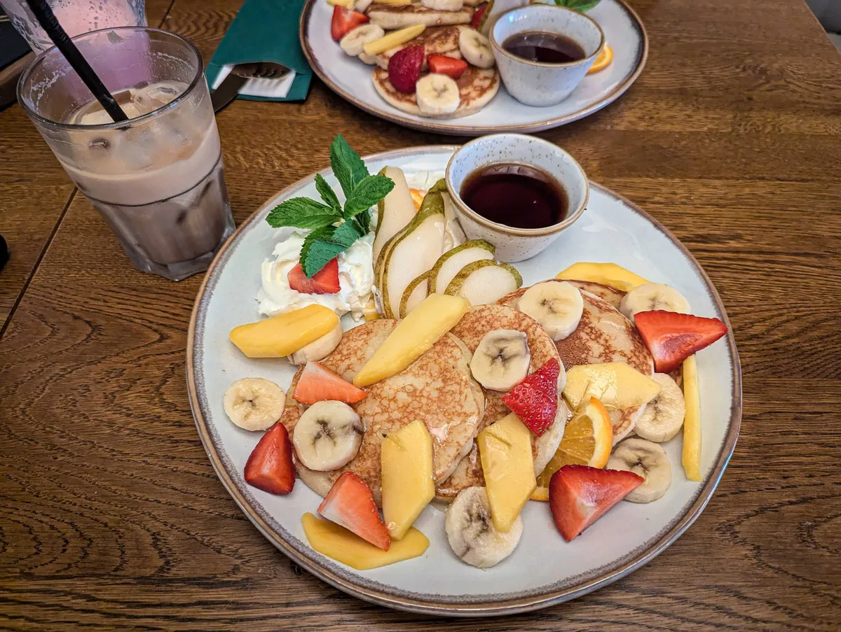 A plate of pancakes with cut strawberries, bananas, mangos, and pears, with whipped cream and maple syrup on the side.