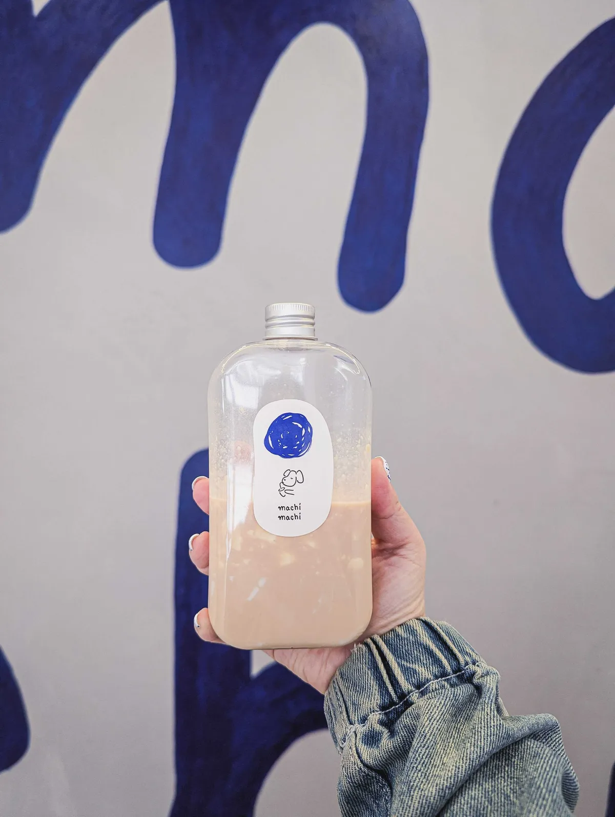 Peige holds a mix of milk tea and panna cotta half-filled inside of a plastic flask in front of a grey concrete wall with large blue letters. There is a white oval sticker with a large blue dot. Below it is the brand's logo, which is a dog drinking bubble tea and 
