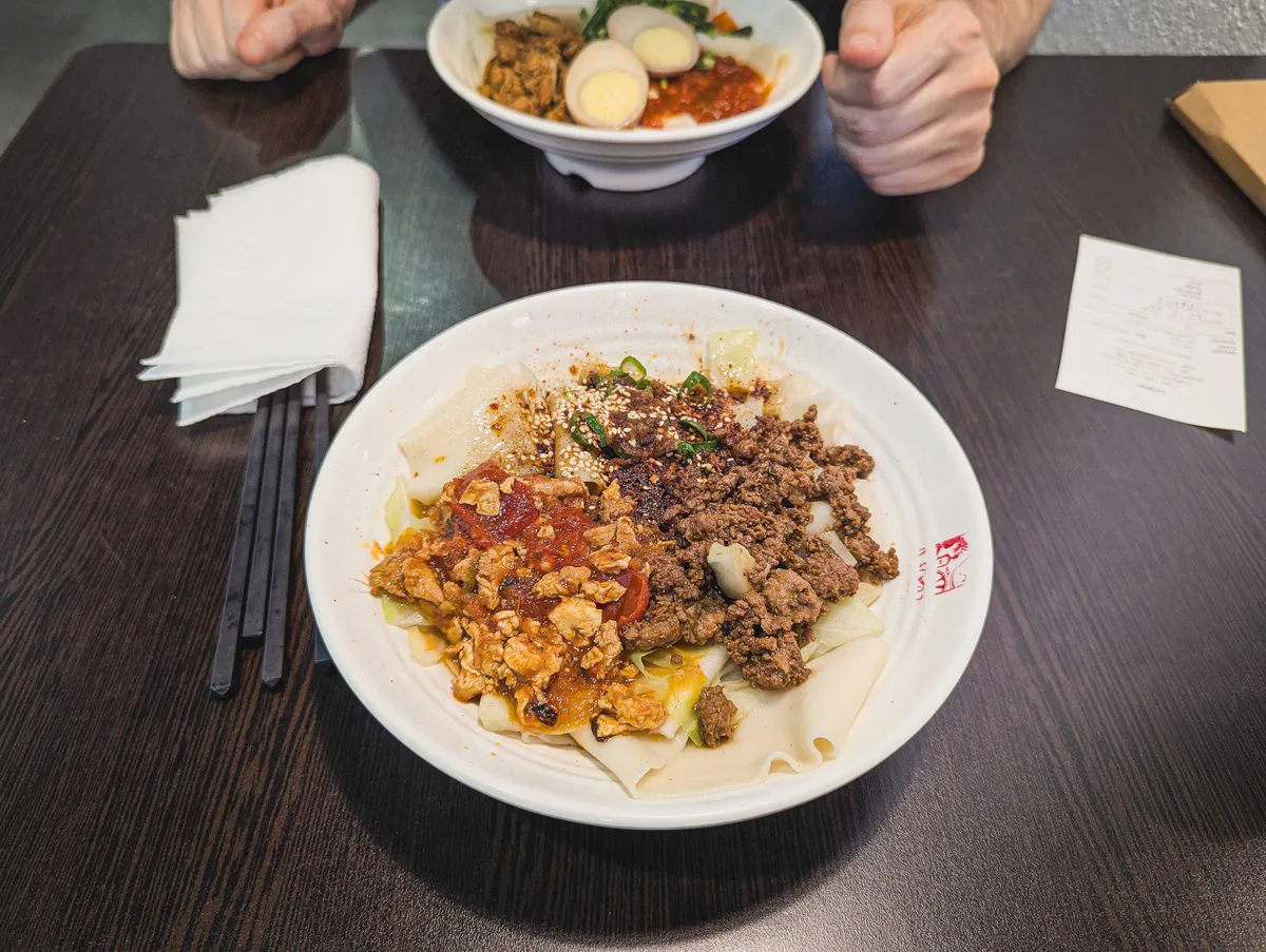A bowl of biang biang noodles topped with scrambled eggs, tomatoes, fried pork, spring onions, chilli flakes, and hot oil.