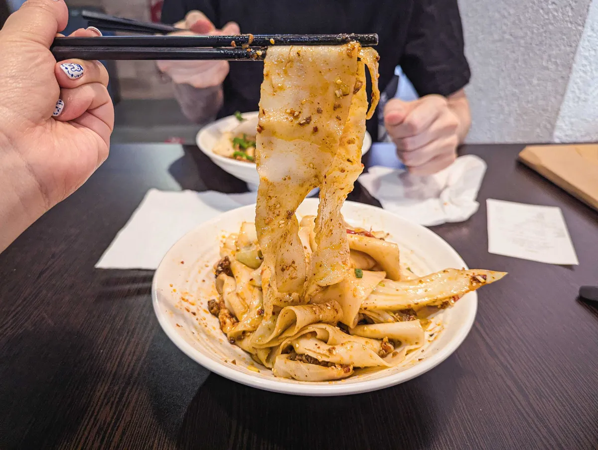 Peige lifting up a flat noodle with their chopsticks.