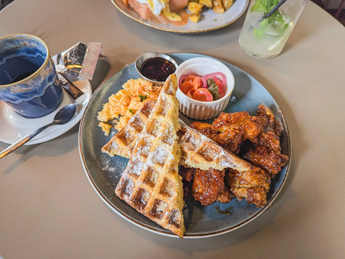 A plate of saucy chicken with triangularly sliced waffles, a small potion of scrambled eggs, and small containers of maple syrup and pickled veggies.