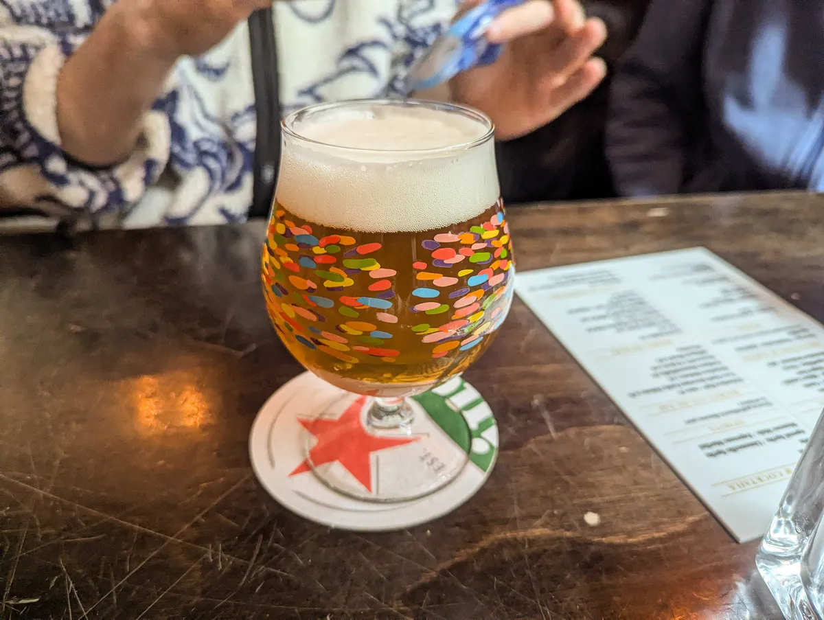 A tulip glass filled with Dutch beer with ample foam. The glass is covered in colorful specks of paint.