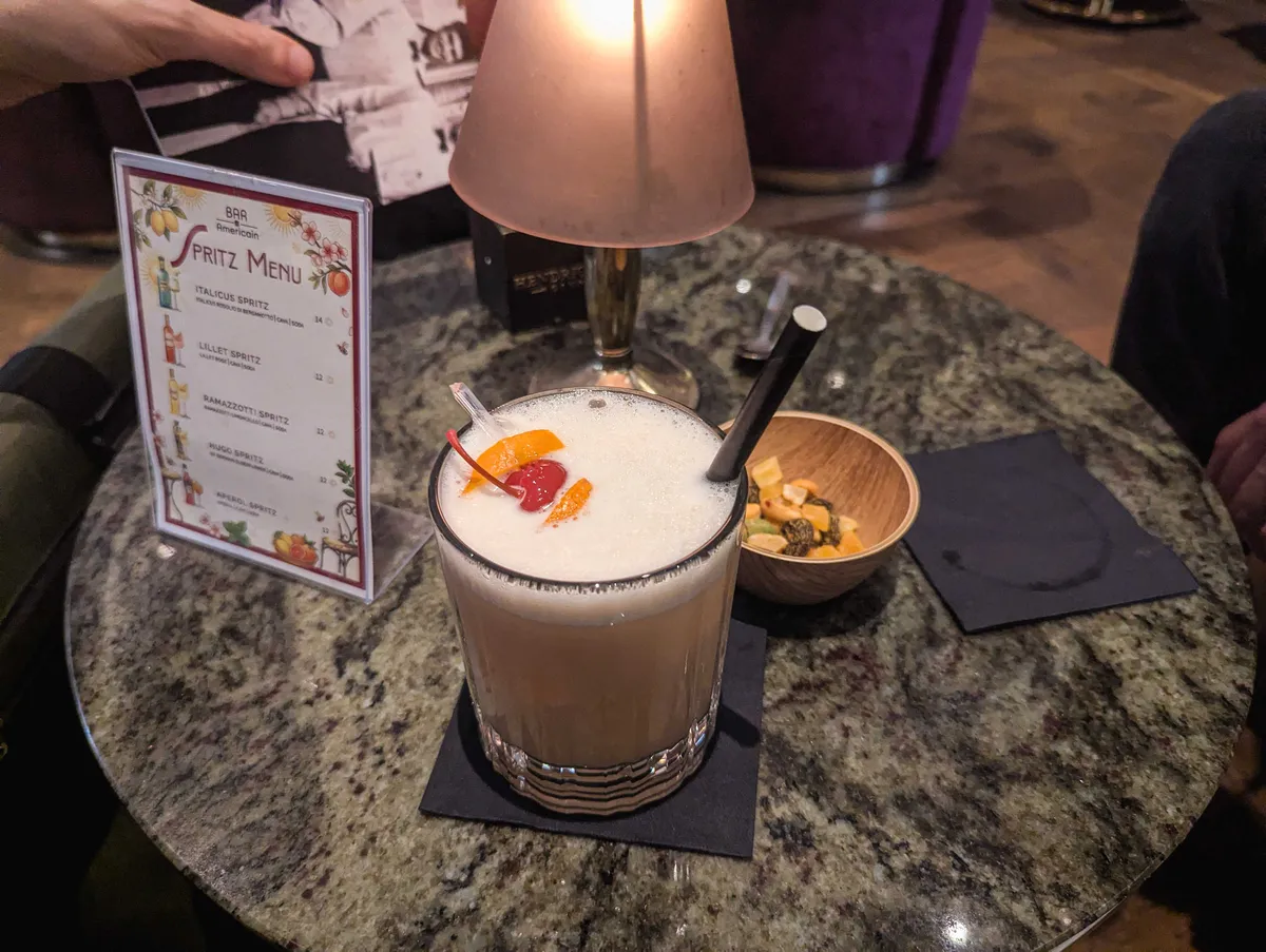 A rocks glass with whiskey sour. There is some foamy egg white and a stick of sliced oranges and a maraschino cherry.