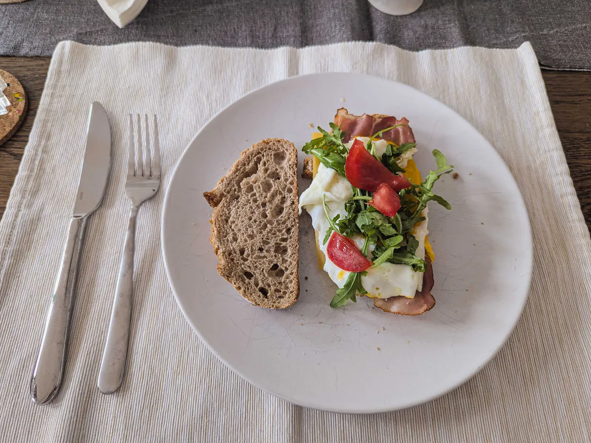 An open-face sandwich on a plate, with bacon, arugula, cherry tomatoes, and a pached egg.
