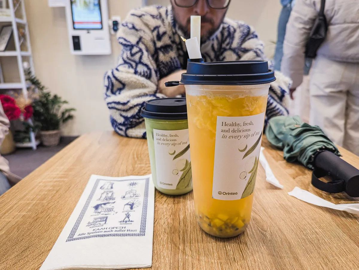 A large plastic takeaway cup of yellow tea with fruit pieces inside. Beside is a shorter cup with a matcha drink.