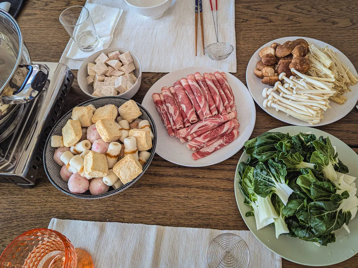 Plates of hot pot ingredients on the other side of the table, including fish balls, taro, beef slices, mushrooms, and bok choy.