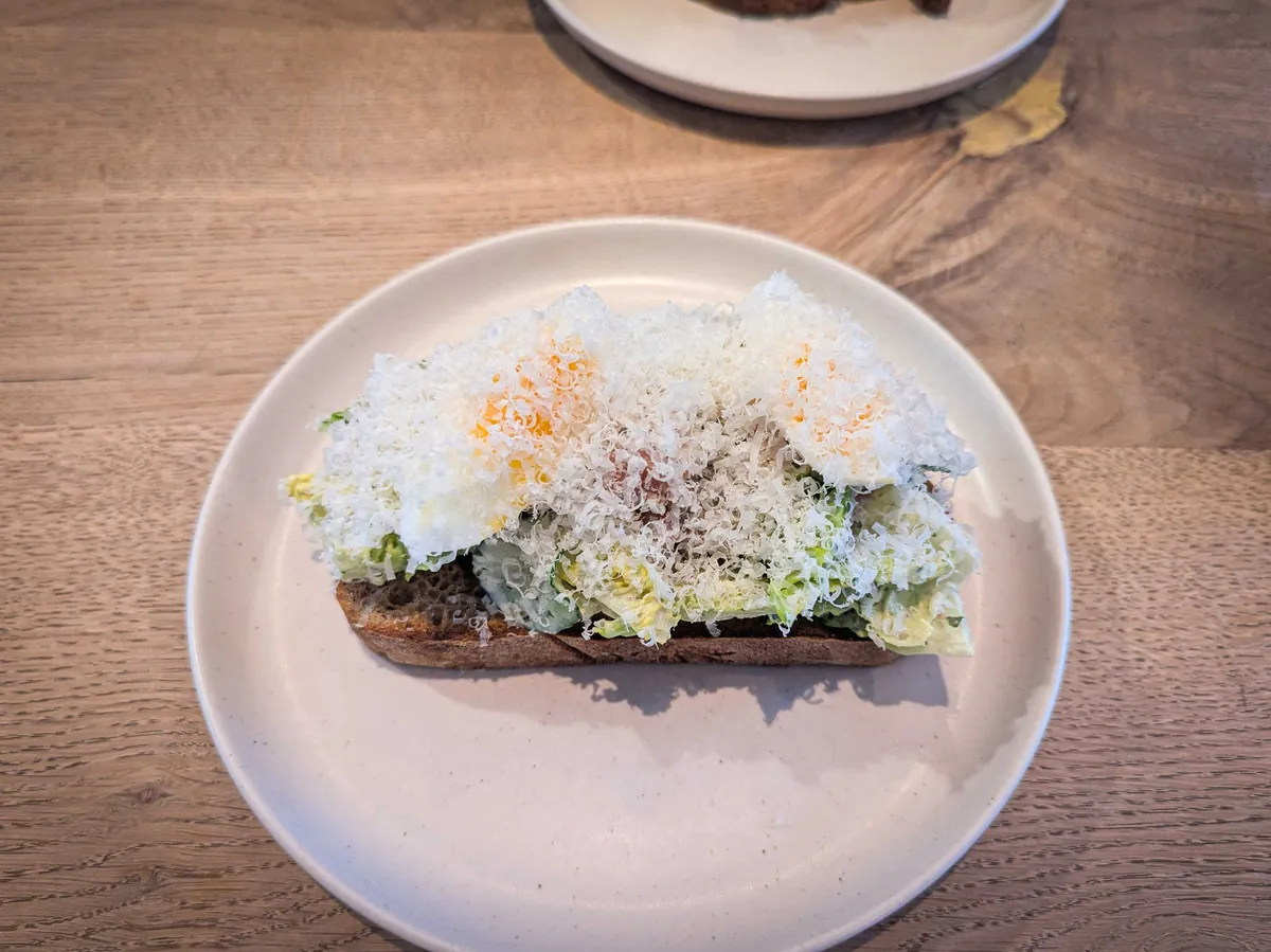 An open-faced toast with avocado, salad, bacon, sliced boiled egg, with a generous amount of finely shredded parmesan on top.