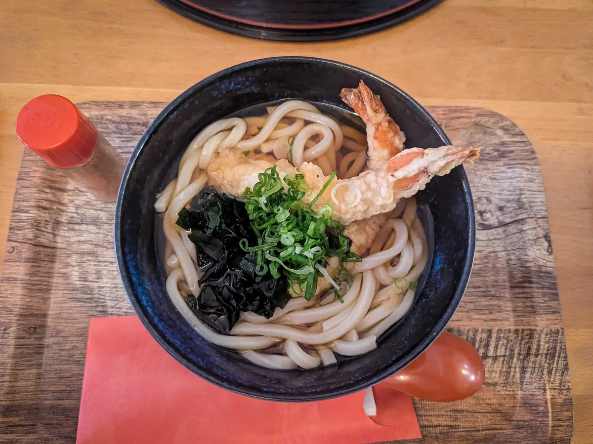 A medium-sized bowl filled with udon and broth, topped with one shrimp tempura, seaweed, and spring onions.