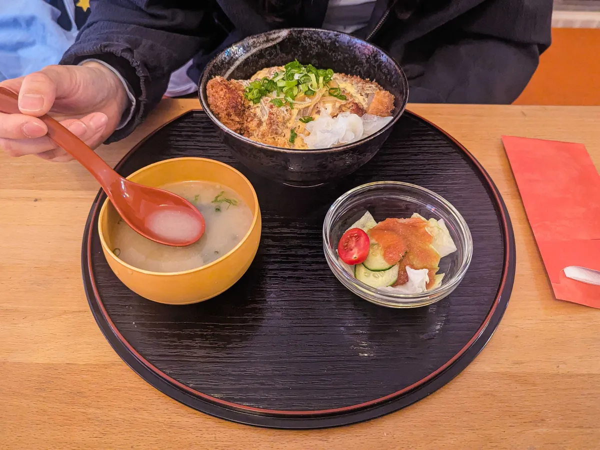 A round tray with a small bowl of miso soup, a small dish of salad, and a medium-sized bowl with rice, katsu, and eggs with spring onions on top.