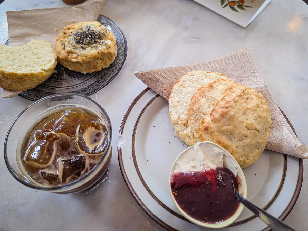 A marble table with two plates of scones. One has seeds on top, the other has a jar of jam and cream next to it. There's an iced coffee in between the plates.
