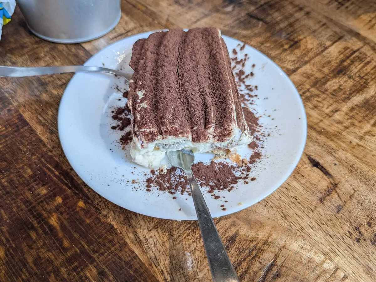 A square of tiramisu on a white plate with two forks underneath the cake.