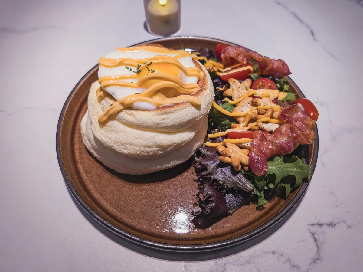A soufflé pancake with a sunny-side-up egg on top, salad and bacon on the side, and hollandaise sauce drizzled over the plate.