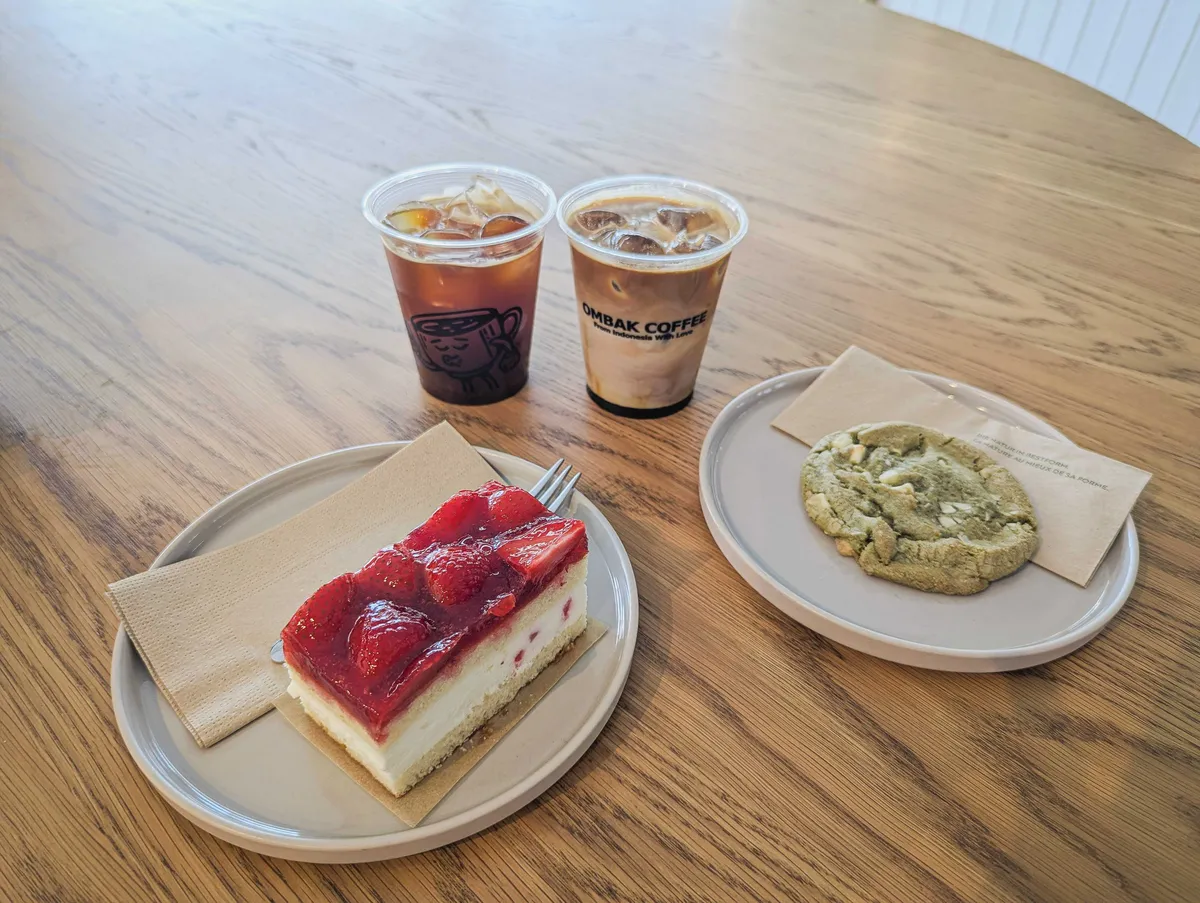 A plate of strawberry shortcake, a plate with a matcha cookie, and two coffee drinks in clear plastic containers on a wooden table.