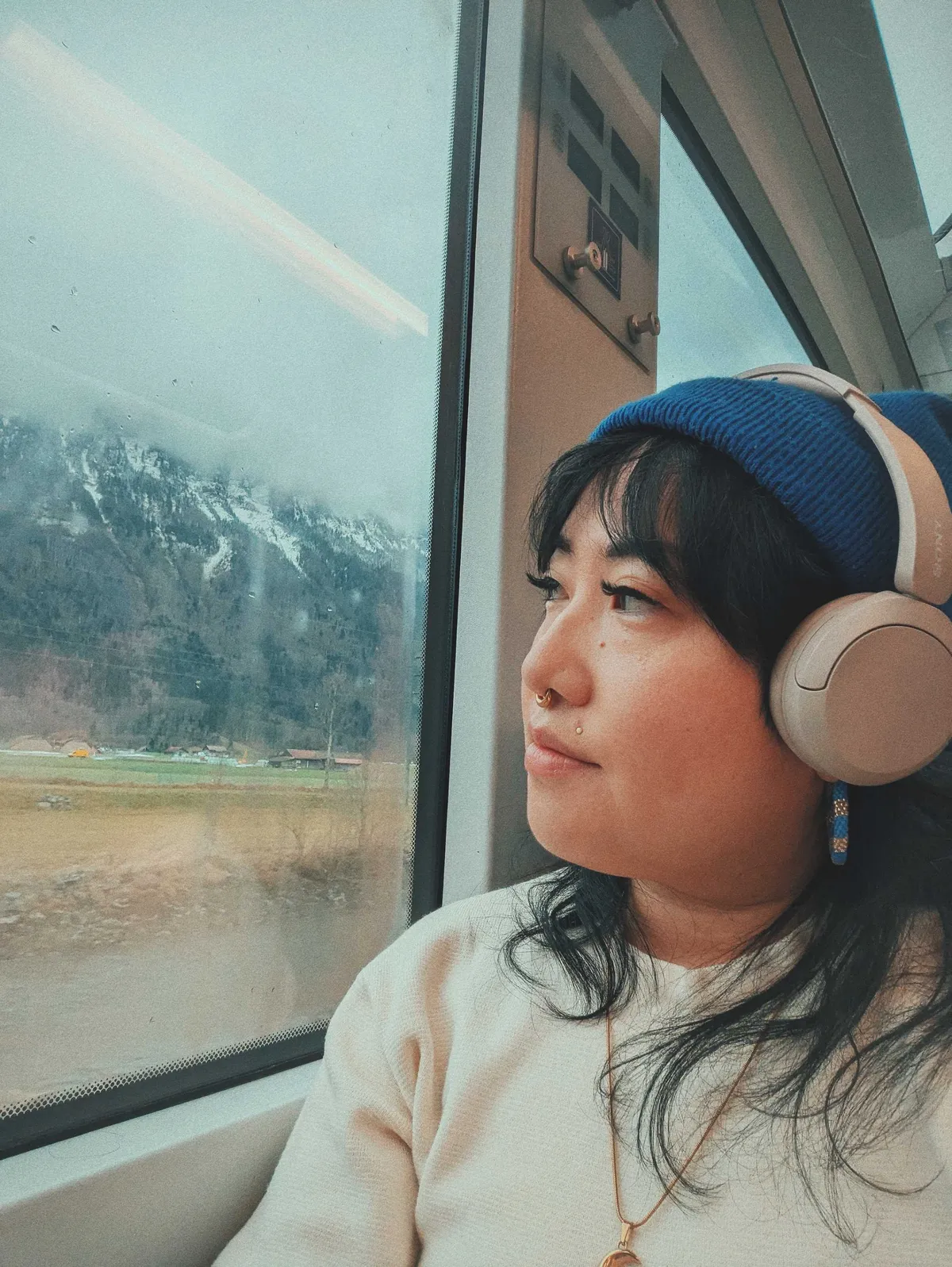 Peige looking pensively out the train window, wearing headphones and a blue beanie.