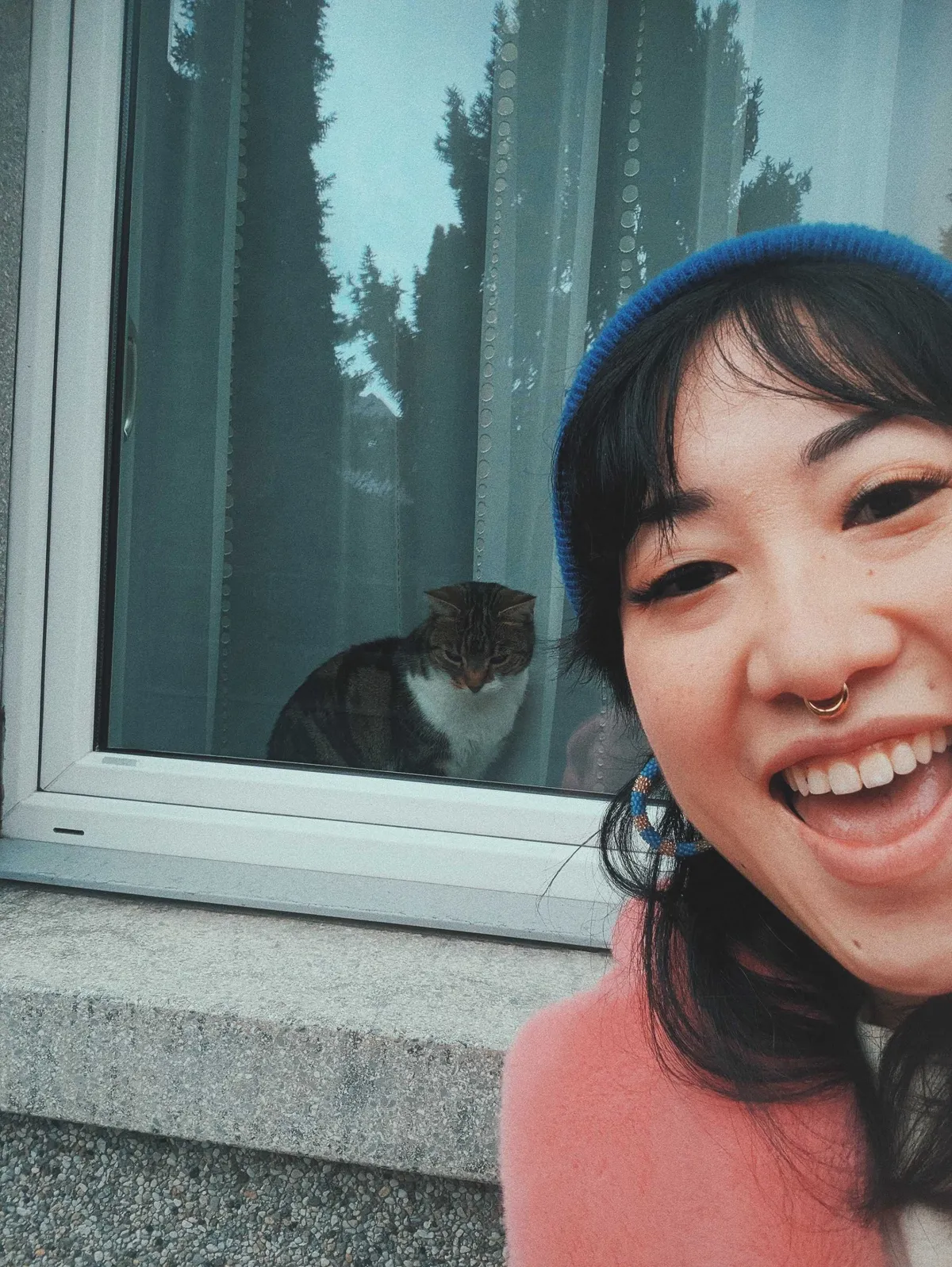 Peige smiling enthusiastically in front of a piebald tabby that's sitting from behind the window pane.