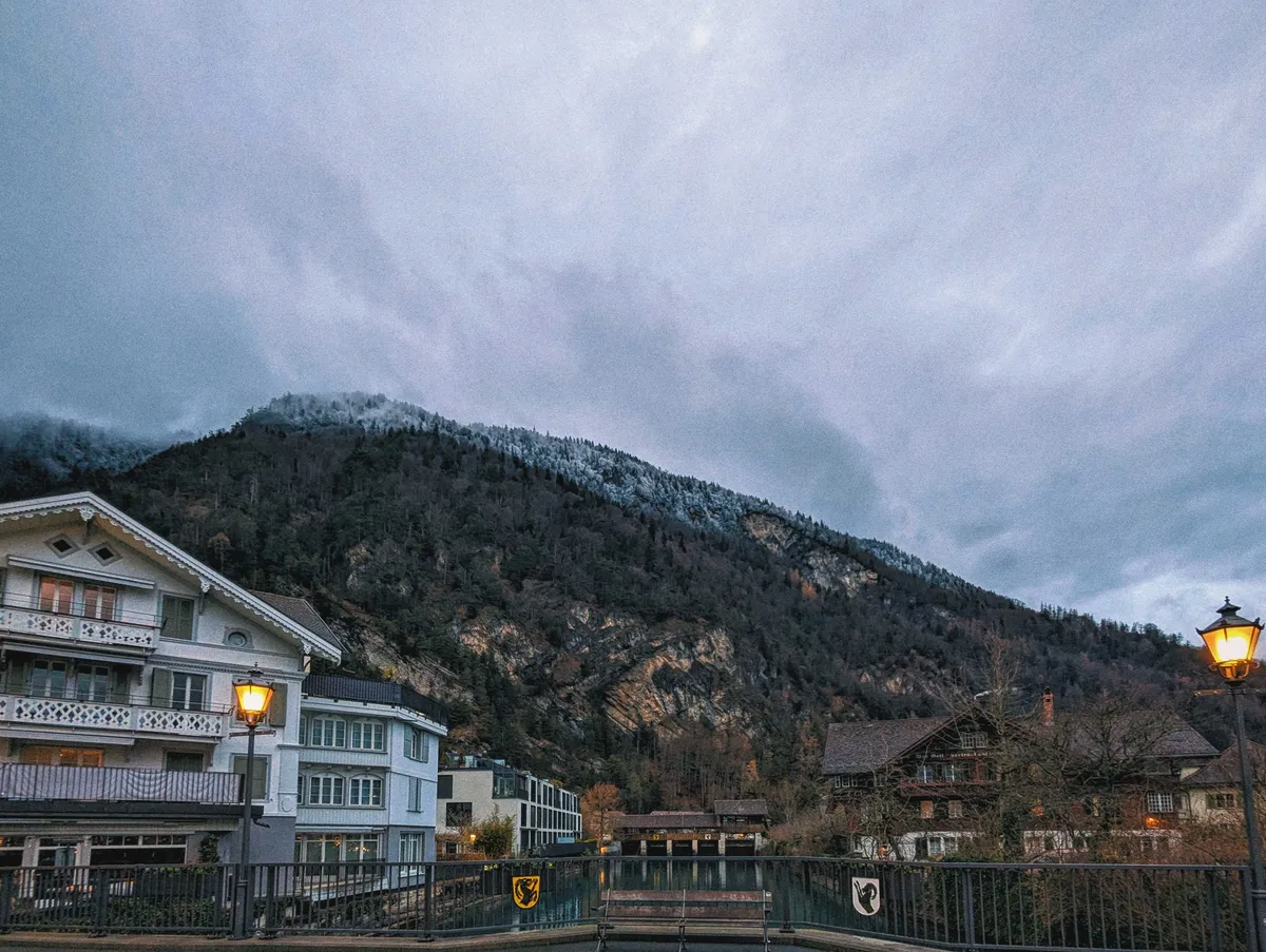 View from the bridge overlooking the river to show the mountain in the background. There are two sigils facing each other, both featuring a black mountain goat. One sigil is golden yellow and the other is white.