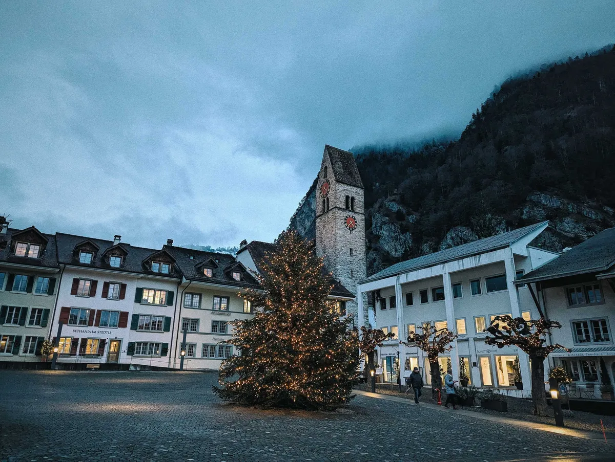 A courtyard with a tall bell tower in the corner. There is a large evergreen tree at the center, decorated with lights. The mountain is in the background and the sky is covered in grey clouds.