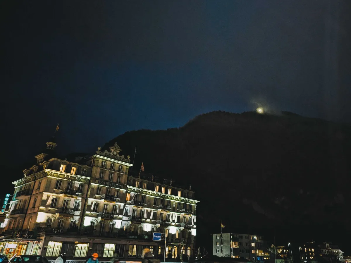 Night time. The mountain is a large, dark shadow. On top is one single bright source of light. There is a hotel in the foreground.