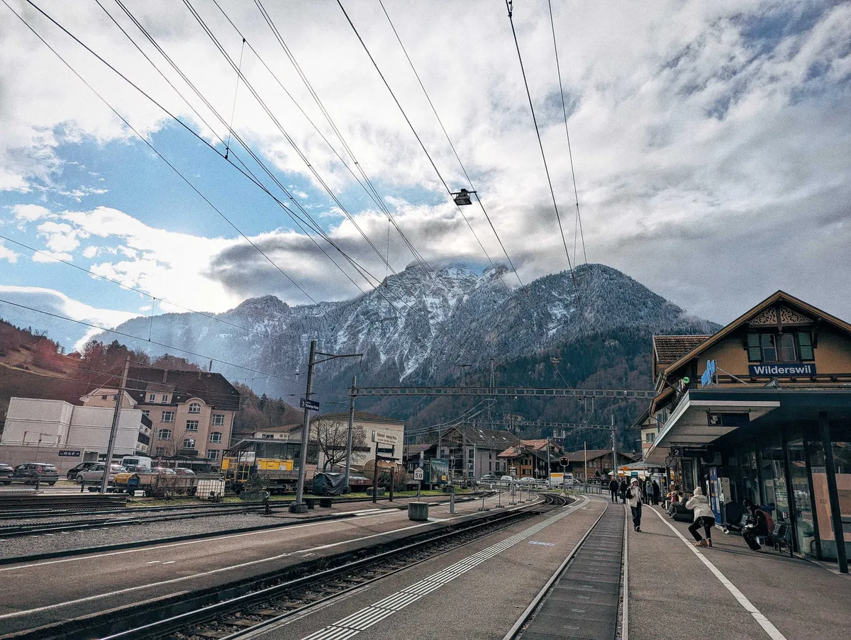 Train tracks leading towards the giant snow-covered mountains, next to a building with a traditional rooftop with a sign that reads, "Wilderswil."