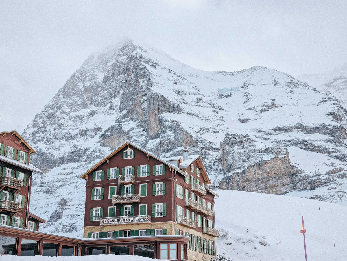 A brown building with green window shutters and the sign, "Des Alpes.". It sits at the foot of a large snow-covered mountain.