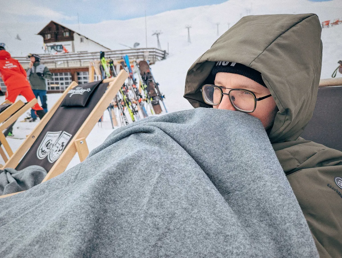A white man wearing glasses huddles under a grey blanket for warmth, covering his mouth.