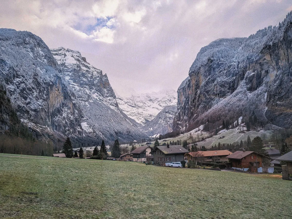 Green fields with houses scattered in the middle. The background opens up to grand, majestic mountains.