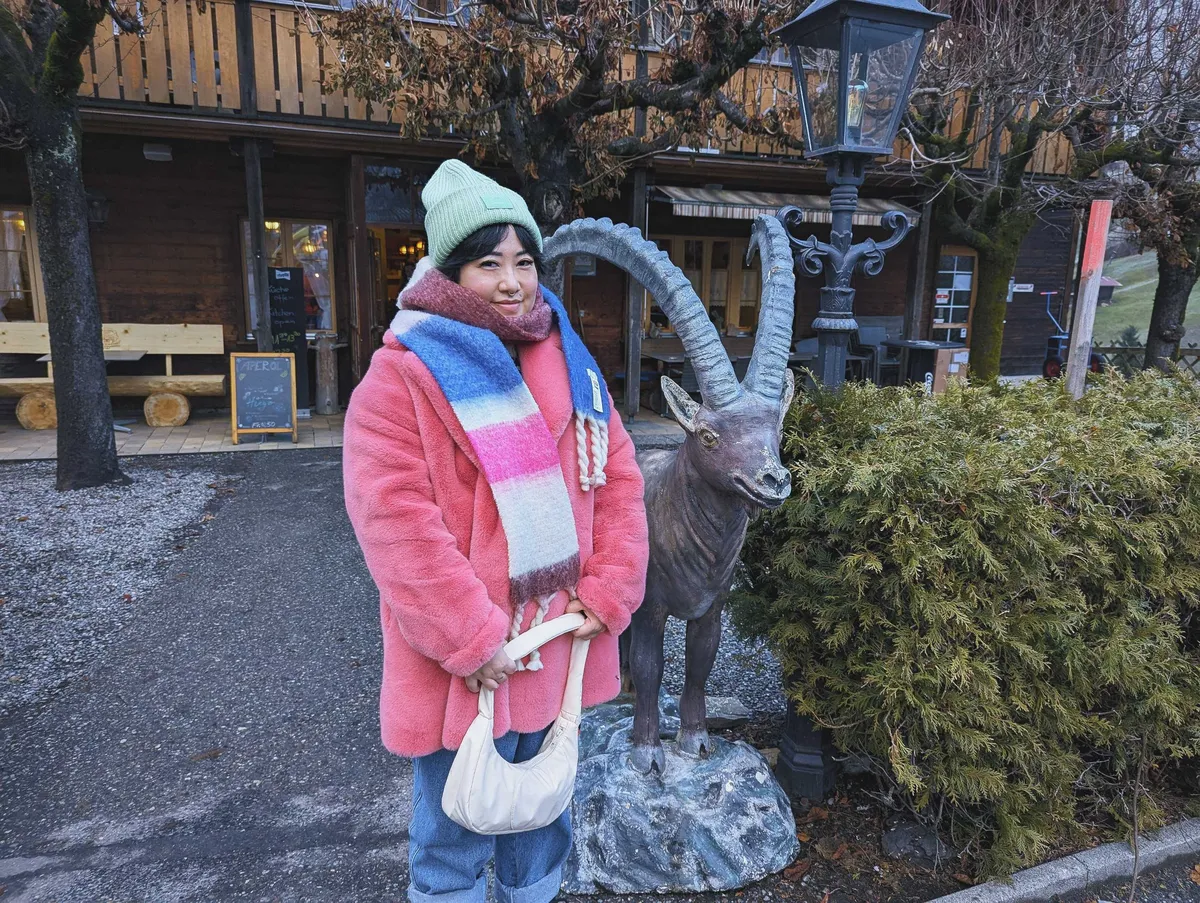 Peige, wearing a pink faux fur coat, colorful scarf, and lime green beanie, poses next to the alpine ibex.