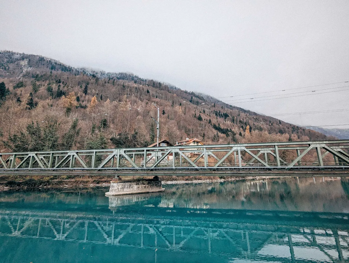 A scenic view of a light green bridge with the foothills in the background, and dark turquoise water below.