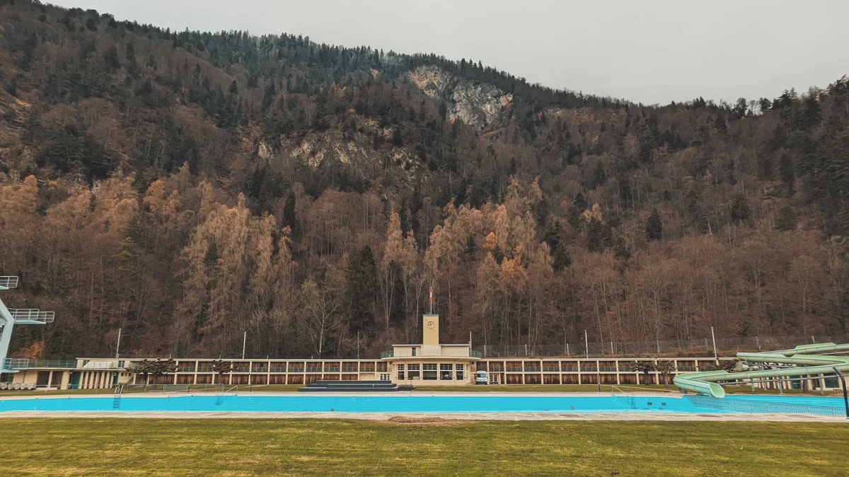 A swimming pool in the foreground with a school-like building in the back (complete with a clock tower). There is a grassy field in front of the pool, and tall brown trees ascending upwards into the mountains in the background.