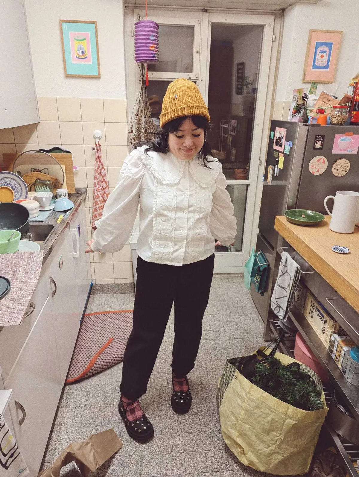 Peige standing in the middle of their messy kitchen, wearing a golden beanie, frilly white blouse, black pants, pink/red checkered wool socks, and black Mary Janes with studs on them.