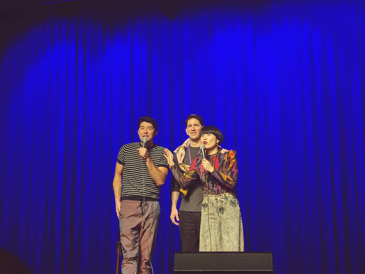 Dylan Adler, Ryan (Atsuko's husband), and Atsuko stand on stage against bright blue curtains.