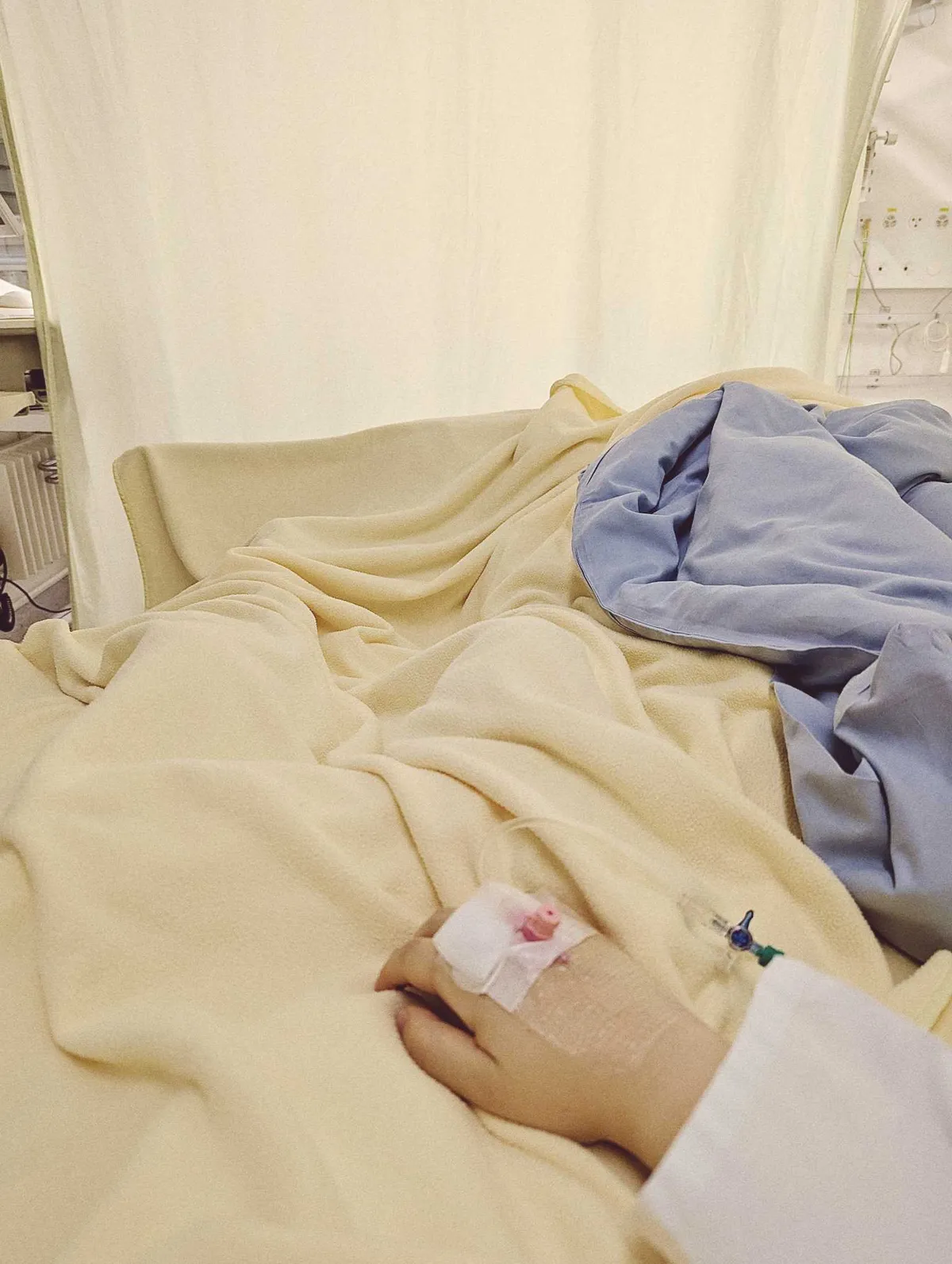 Peige's hand with a pink IV sticking out, lying on cream and light blue hospital blankets.