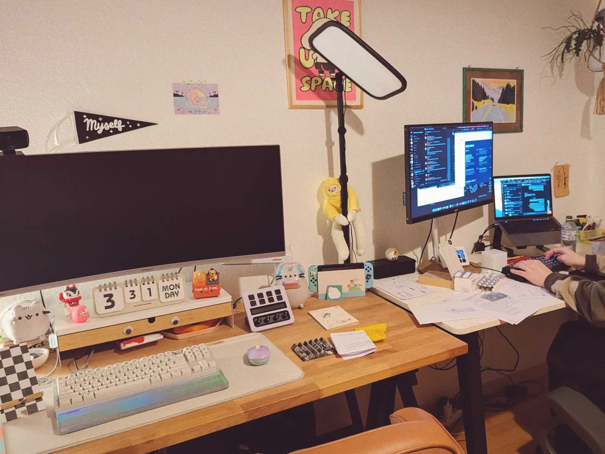 Two desks with large monitors are placed side-by-side. One desk is decorated with cute figurines. The other is stacked with papers, with Daniel's hands at the edge of the photo.
