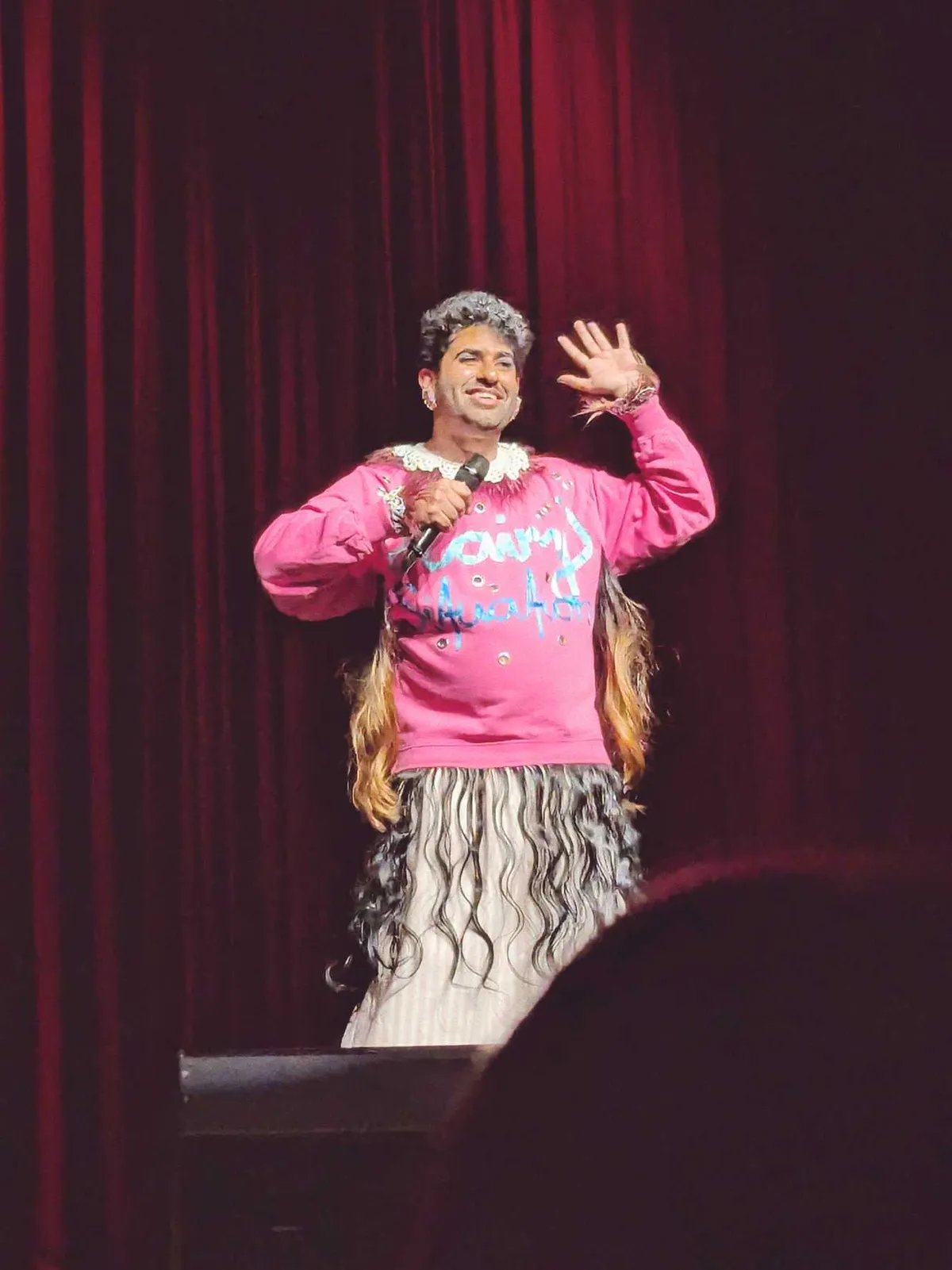 Alok stands with one hand in the air, mid-speech. They are wearing a bright pink sweater with wavy extensions sewn on.