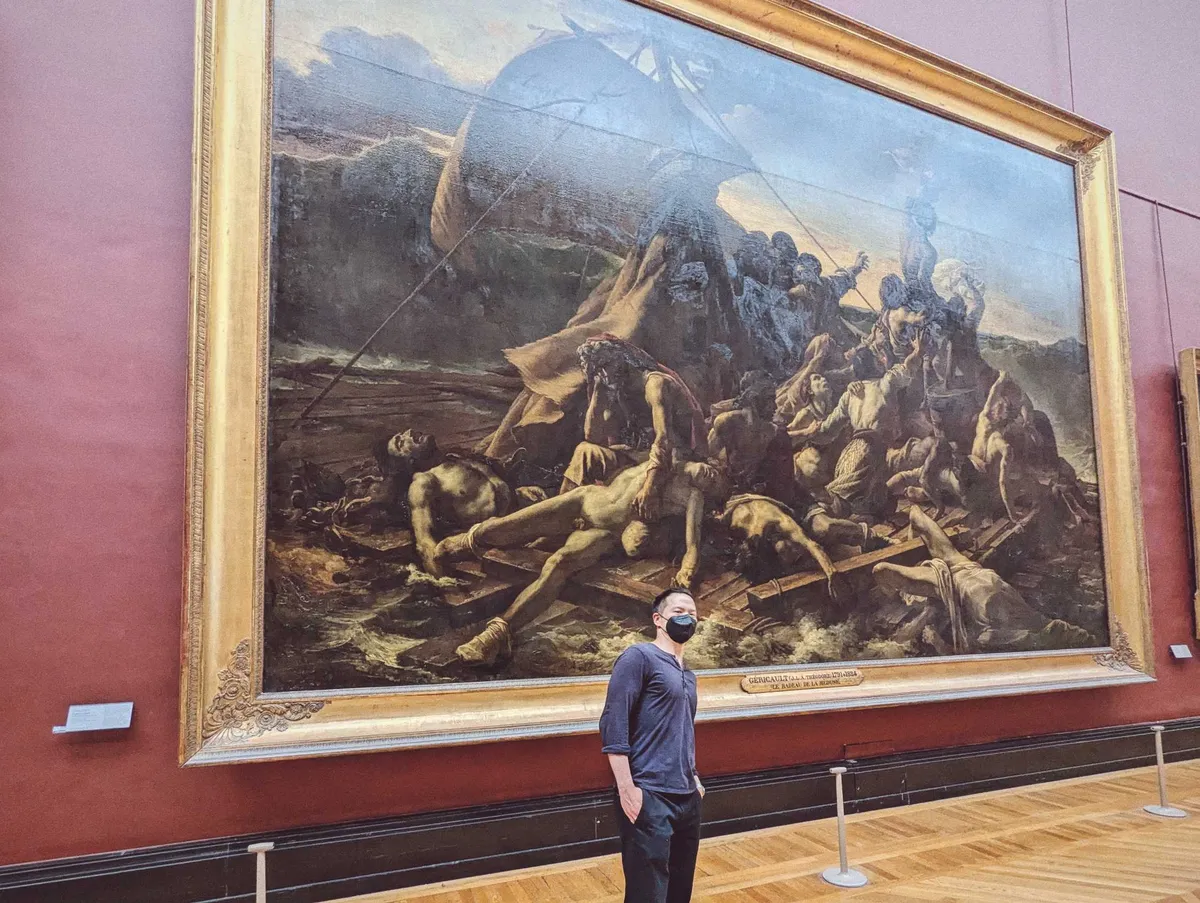 Pat stands in front of a large painting, wearing a black face mask.