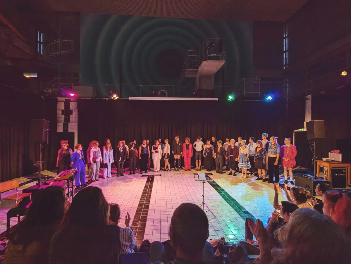 An ensemble of fabulously-dressed choir singers stand in a semi-circle, receiving their applause from the audience.