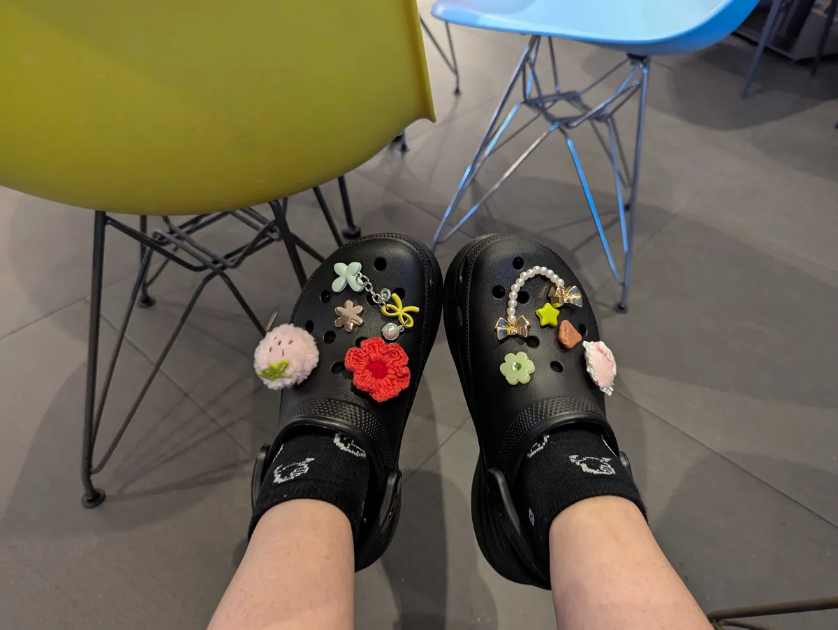 A pair of black crocs with a colorful array of jibbitz attached to them, such as a fuzzy pink strawberry, lots of colorful flowers, and a string of fake pearls.